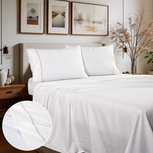 May include: White bed sheet set with two pillowcases. The pillowcases and the edge of the flat sheet have a delicate, light green embroidered design. The bedding is neatly arranged on a bed, with a close-up view of the embroidered detail.