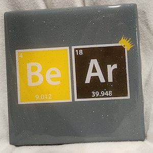 May include: A gray square tile with a yellow and black square on it. The yellow square has the number 4 and the letters "Be" on it. The black square has the number 18 and the letters "Ar" on it. The tile has a small gold crown on the top right corner.