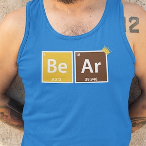 以下が含まれることがあります： 周期表にインスパイアされたデザインの青いタンクトップ。「Be Ar」の文字が黄色と茶色のボックスで表示され、「Ar」の上に小さな王冠があります。タンクトップは柔らかい素材で作られています。