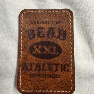 Puede incluir: Un parche rectangular de cuero marrón con costuras blancas. El parche presenta las palabras "PROPERTY OF BEAR XXL ATHLETIC DEPARTMENT" en una fuente marrón oscuro.