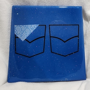 Op de afbeelding: Blauwe vierkante onderzetter met een glinsterende afwerking en twee zwarte gecontourde jean-zakken. De bovenste zak heeft een wit paisley bandana-patroon.