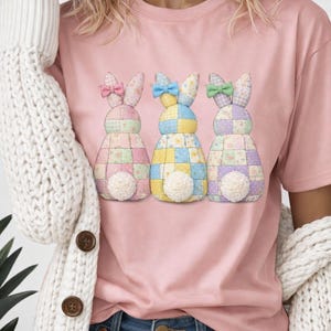 Pode incluir: Camiseta rosa com três desenhos de coelhos de patchwork com padrões coloridos, laços e caudas brancas e fofas. Os coelhos estão dispostos em fila, adicionando um toque de fantasia ao design da camiseta.