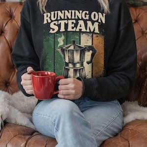 Puede incluir: Sudadera negra con el texto "RUNNING ON STEAM" sobre un gráfico de cafetera. El gráfico tiene rayas verdes, blancas y naranjas. Una persona sostiene una taza roja mientras está sentada en un sofá de cuero marrón.