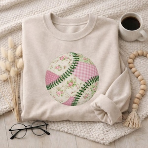Puede incluir: Sudadera beige con un diseño de béisbol con parches florales y de cuadros rosas y blancos, cosidos con hilo verde. La sudadera está doblada sobre una superficie texturizada de color crema, con una taza de café y cuentas decorativas cerca.