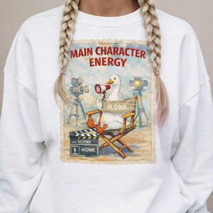 May include: White sweatshirt with a graphic featuring a duck in a director's chair holding a megaphone. The text "MAIN CHARACTER ENERGY" is above the duck, with "H.ONK" on the chair. A film clapperboard reads "SCENE 1 HONK".