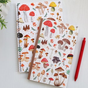 May include: Two spiral bound notebooks with a white background and a colorful mushroom pattern. The notebooks are on a white surface with a red pen.