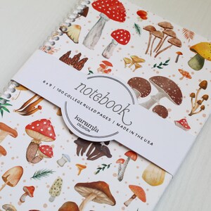 May include: A white notebook with a mushroom pattern. The cover features a variety of mushrooms in different colors and sizes, including red, brown, and yellow. The notebook is 6 x 9 inches and has 100 college ruled pages. The text "notebook" is written in a cursive font on the cover, along with the words "karamia design" and "made in the USA".