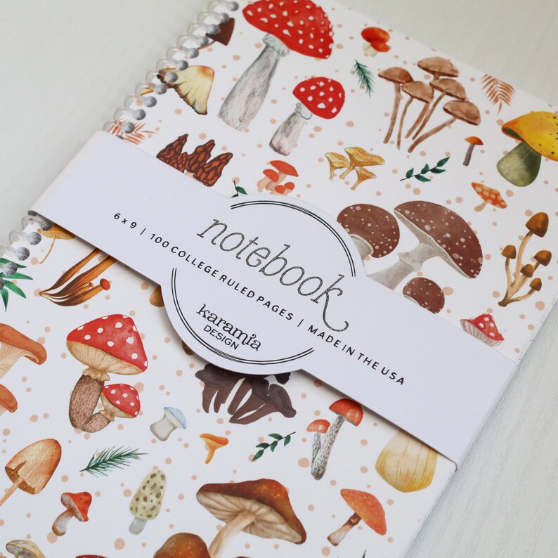 May include: A white notebook with a mushroom pattern. The cover features a variety of mushrooms in different colors and sizes, including red, brown, and yellow. The notebook is 6 x 9 inches and has 100 college ruled pages. The text "notebook" is written in a cursive font on the cover, along with the words "karamia design" and "made in the USA".