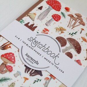 May include: A white sketchbook with a brown spine and a mushroom pattern. The cover has a sticker with the text "sketchbook" in a circle, "5 x 8 | 100 BLANK PAGES | MADE IN THE USA" and "karamia DESIGN".