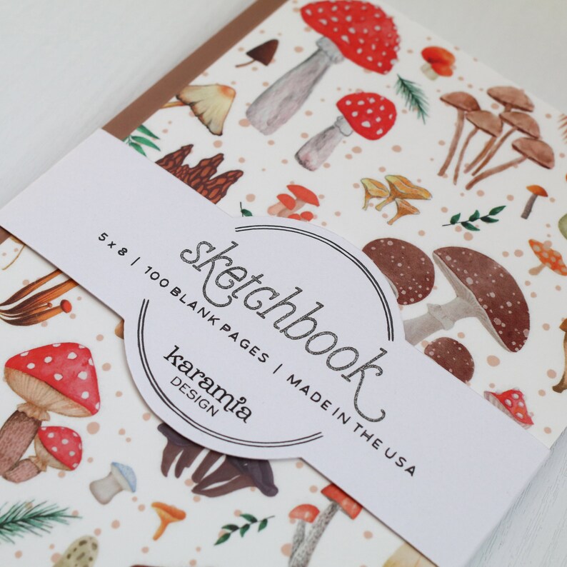 May include: A white sketchbook with a brown spine and a mushroom pattern. The cover has a sticker with the text "sketchbook" in a circle, "5 x 8 | 100 BLANK PAGES | MADE IN THE USA" and "karamia DESIGN".