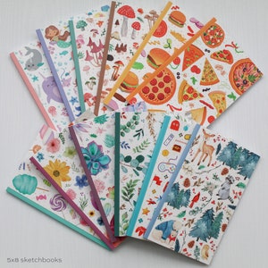 May include: A set of eight 5x8 inch sketchbooks with various colorful and whimsical designs. The designs include mushrooms, pizza, narwhals, succulents, and a winter forest scene.