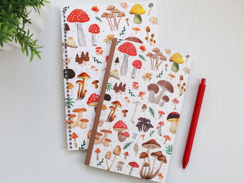 May include: Two spiral bound notebooks with a white background and a colorful mushroom pattern. The notebooks are on a white surface with a red pen.