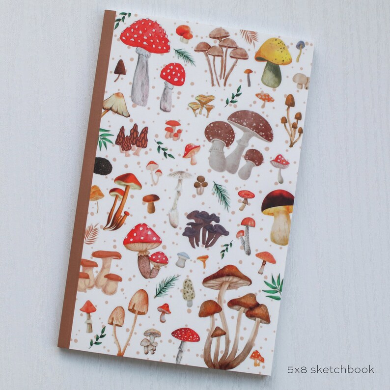 May include: A white sketchbook with a brown spine and a colorful mushroom pattern. The mushrooms are red, orange, yellow, brown, and white. The sketchbook is 5x8 inches.