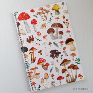 May include: A white spiral bound notebook with a colorful mushroom pattern. The cover features red, brown, yellow, and white mushrooms on a white background with small white dots. The text "6x9 notebook" is printed in black at the bottom of the cover.