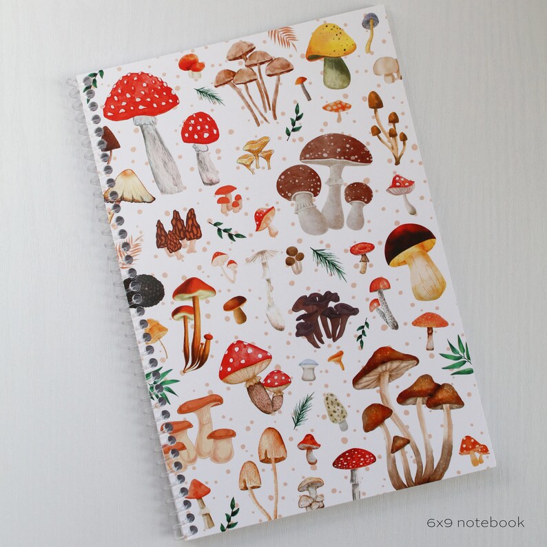 May include: A white spiral bound notebook with a colorful mushroom pattern. The cover features red, brown, yellow, and white mushrooms on a white background with small white dots. The text "6x9 notebook" is printed in black at the bottom of the cover.
