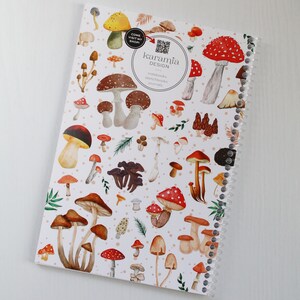 May include: A white spiral bound notebook with a colorful mushroom design. The cover features red and white mushrooms, brown mushrooms, and green leaves. The text "karamia DESIGN" is printed on the cover.