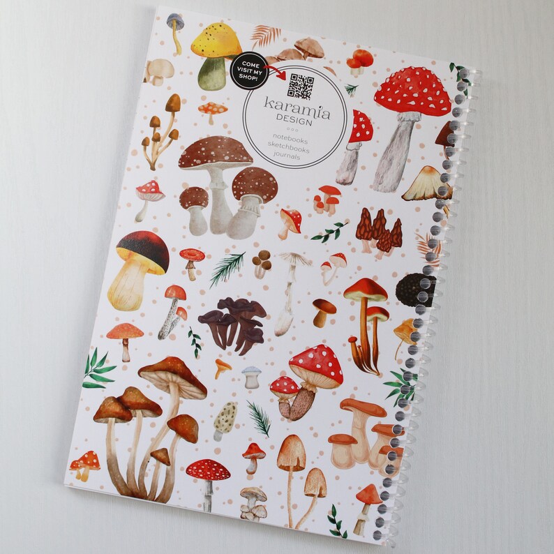 May include: A white spiral bound notebook with a colorful mushroom design. The cover features red and white mushrooms, brown mushrooms, and green leaves. The text "karamia DESIGN" is printed on the cover.