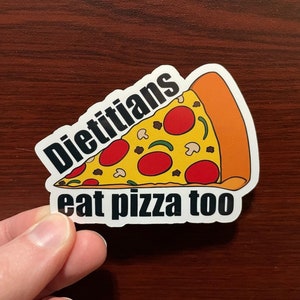 May include: A white sticker with a black outline of a pizza slice with red pepperoni, green peppers, and mushrooms. The text "Dietitians eat pizza too" is written in black below the pizza slice.