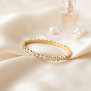 May include: A gold-toned hinged bangle bracelet featuring a continuous row of round, clear, bezel-set stones. The bracelet has a polished metallic finish and a secure clasp mechanism, showcasing a uniform design of sparkling circular accents.
