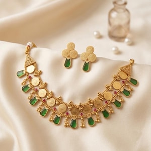 May include: A gold-toned necklace and matching stud earrings set featuring circular coin-like charms, teardrop-shaped green stones, and small red accents. The necklace has a multi-layered chain design with intricate metalwork and small white beads.