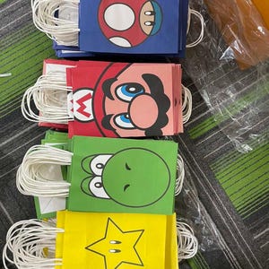 May include: A collection of colourful paper gift bags featuring characters from a popular video game. The bags are blue, red, green, and yellow, with white handles. Each bag has a different character design, including a mushroom, a character's face, a green dinosaur, and a star.