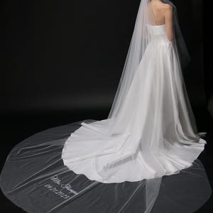 Può includere: Abito da sposa bianco senza spalline con lungo strascico e velo trasparente. Il velo è ricamato con la scritta "Mrs. Thompson 09.21.2025". L'abito ha un corpetto aderente e una gonna fluida, su sfondo nero.