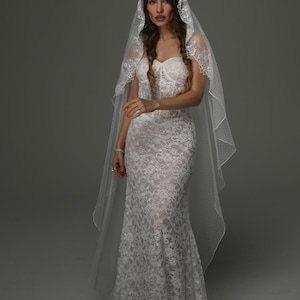 May include: A white lace wedding dress with a mermaid silhouette. The dress has a sweetheart neckline and is worn with a matching lace veil. The bride is wearing a bracelet and ring.