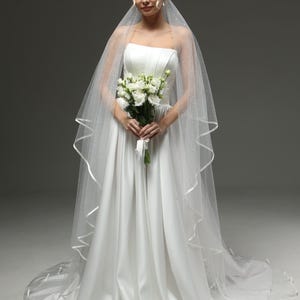 Op de afbeelding: Een witte bruidsjurk met een strapless lijfje en een zwierige rok. De jurk wordt gecombineerd met een witte sluier met een gegolfde rand.
