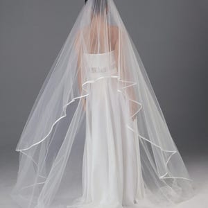May include: A long, white wedding veil with a ribbon edge, worn over a white bridal gown. The veil cascades down, adding a traditional touch. The dress has a simple, elegant design.