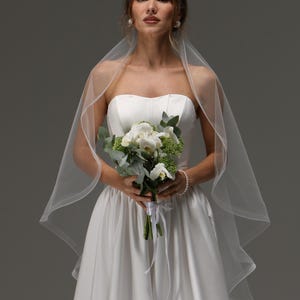 May include: A bride in a white strapless wedding dress and veil holds a bouquet of white flowers and greenery. The dress has a fitted bodice and a flowing skirt. The veil is sheer and drapes over the shoulders. The bouquet is tied with white ribbon.