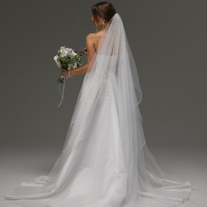 May include: A white wedding dress with a long train and a sheer veil. The bride holds a bouquet of white flowers with green accents. The veil flows down the back of the gown.
