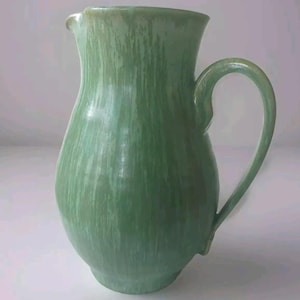 Op de afbeelding: Een groene keramische kan met een getextureerd, gestreept glazuur. De kan heeft een brede basis en een rond lichaam, met een handvat dat omhoog en over de bovenkant buigt.