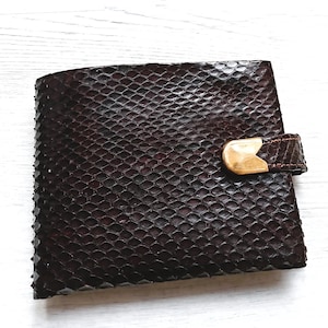 Könnte beinhalten: Eine dunkelbraune Geldbörse mit einem strukturierten, schuppenartigen Muster. Die Geldbörse hat einen Klappenverschluss mit einem braunen Riemen und einem goldfarbenen Metallverschluss. Die Geldbörse ist rechteckig und scheint aus Leder zu sein.