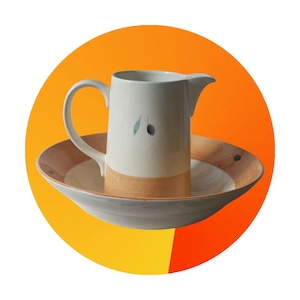 Puede incluir: Una jarra de cerámica blanca con un borde marrón y un diseño de rama de olivo se asienta sobre un platillo marrón y blanco. La jarra tiene un asa y un pico.