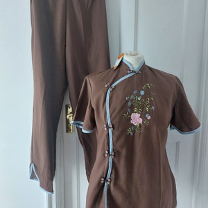 Op de afbeelding: Een bruine twee-delige outfit met een top met mandarijnkraag en een broek. De top heeft een blauwe bies en bloemenborduurwerk. De broek heeft een blauwe bies aan de onderkant.