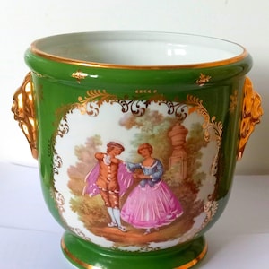 Op de afbeelding: Een groene keramische plantenbak met gouden details en handvatten. De plantenbak heeft een geschilderde scène van een stel in een tuin. De rand en de basis zijn afgewerkt met goud. Een decoratief item.