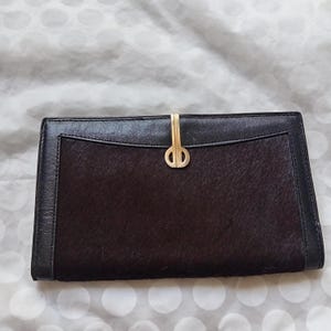 Puede incluir: Una cartera rectangular de cuero negro con un panel central marrón oscuro y texturizado. La cartera presenta un cierre y un ribete dorados. El diseño es sencillo y elegante, adecuado para el uso diario.