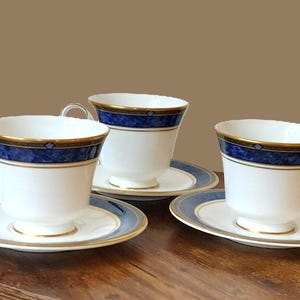 May include: Three white teacups and saucers with a blue and gold decorative band. The teacups have curved handles and sit on matching saucers. The set is arranged on a wooden surface.
