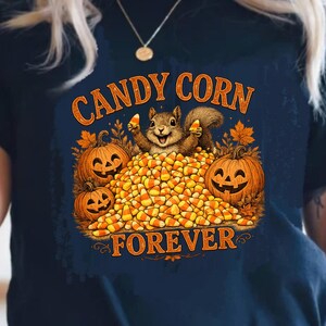 May include: A dark navy blue t-shirt featuring a graphic print of a squirrel sitting on a large pile of candy corn, flanked by two jack-o'-lanterns. The text "CANDY CORN FOREVER" is printed in orange above and below the central illustration.