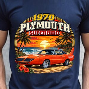 May include: A navy blue t-shirt featuring a graphic print of an orange 1970 Plymouth Superbird car on a beach at sunset. The text reads 1970 PLYMOUTH SUPERBIRD. The design includes palm trees, ocean waves, and red hibiscus flowers at the bottom.