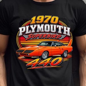 May include: A black short-sleeve t-shirt featuring a graphic of an orange 1970 Plymouth Superbird car. The design includes the text 1970 PLYMOUTH SUPERBIRD and 440 in bold lettering set against a circular yellow and orange sunset background.