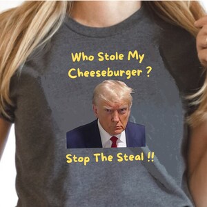 May include: A dark gray t-shirt with a yellow graphic of a man's face and the text "Who Stole My Cheeseburger? Stop The Steal!!"