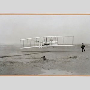 May include: A black and white photograph of the Wright brothers' first successful aeroplane flight. The plane is in the air, and a man is standing on the ground watching it.