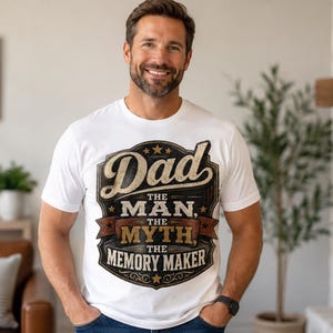 Pode incluir: Uma t-shirt branca de manga curta com um grande gráfico de estilo vintage no peito. O design inclui o texto "Dad THE MAN, THE MYTH, THE MEMORY MAKER" num emblema decorativo em camadas com tons de castanho, preto e creme.