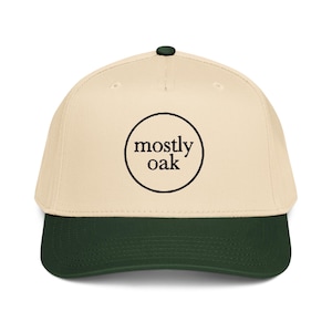 May include: A baseball cap with a cream-colored crown and a dark green brim. The cap features the text "mostly oak" in a black circular design on the front. The cap has a dark green button on top.