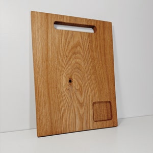 May include: A rectangular wooden cutting board with a handle cutout and a small rectangular recess. The board has a natural wood grain pattern and is light brown in color.