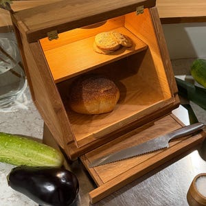 May include: A wooden bread box with a hinged lid and interior lighting. The box features two shelves holding a round loaf of bread and two cookies. A serrated bread knife rests on a wooden cutting board base attached to the front of the unit.