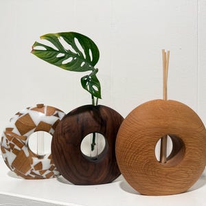 May include: Three decorative wooden vases with circular openings. One vase is a mosaic of wood and white resin, another is dark brown, and the third is light brown. A green plant with a unique leaf shape is in the dark brown vase.