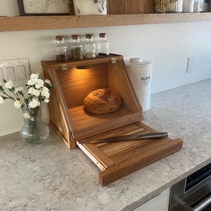 May include: A wooden bread box with a glass door and a pull-out cutting board. The bread box is on a countertop with a loaf of bread inside. There are four small glass jars with lids on the shelf above the bread box.