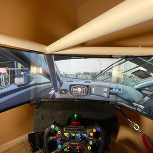 Op de afbeelding: Een race-simulatoropstelling met drie gebogen computermonitoren die een autoracespel weergeven. Een zwart racestuur met kleurrijke knoppen en een zwarte basis staan op de voorgrond. De monitoren zijn op een zwarte standaard gemonteerd.